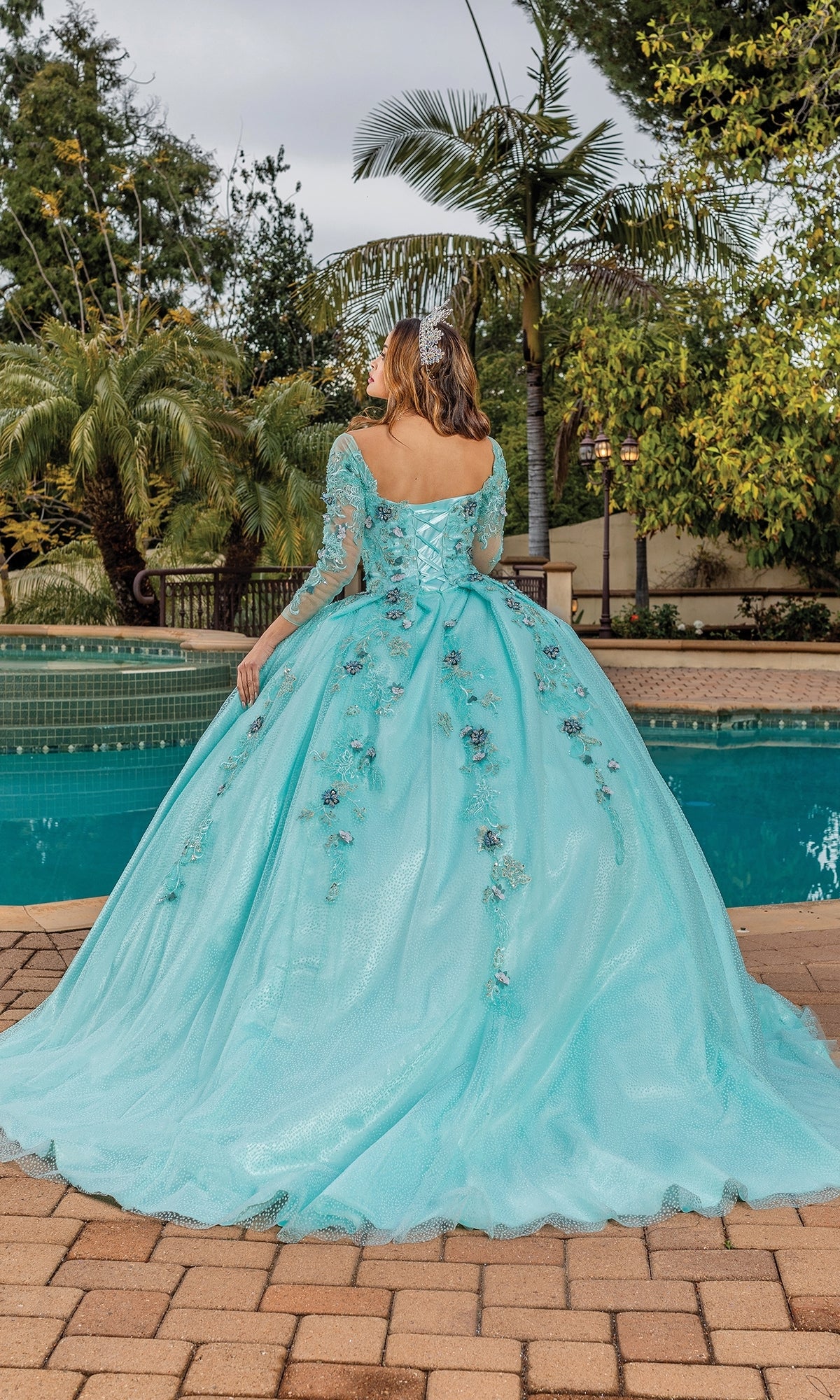A woman stands outdoors by a pool, facing away, wearing the Quinceanera Dress 1847 By Dancing Queen in mint green with floral details and long sheer sleeves, with trees visible in the background.